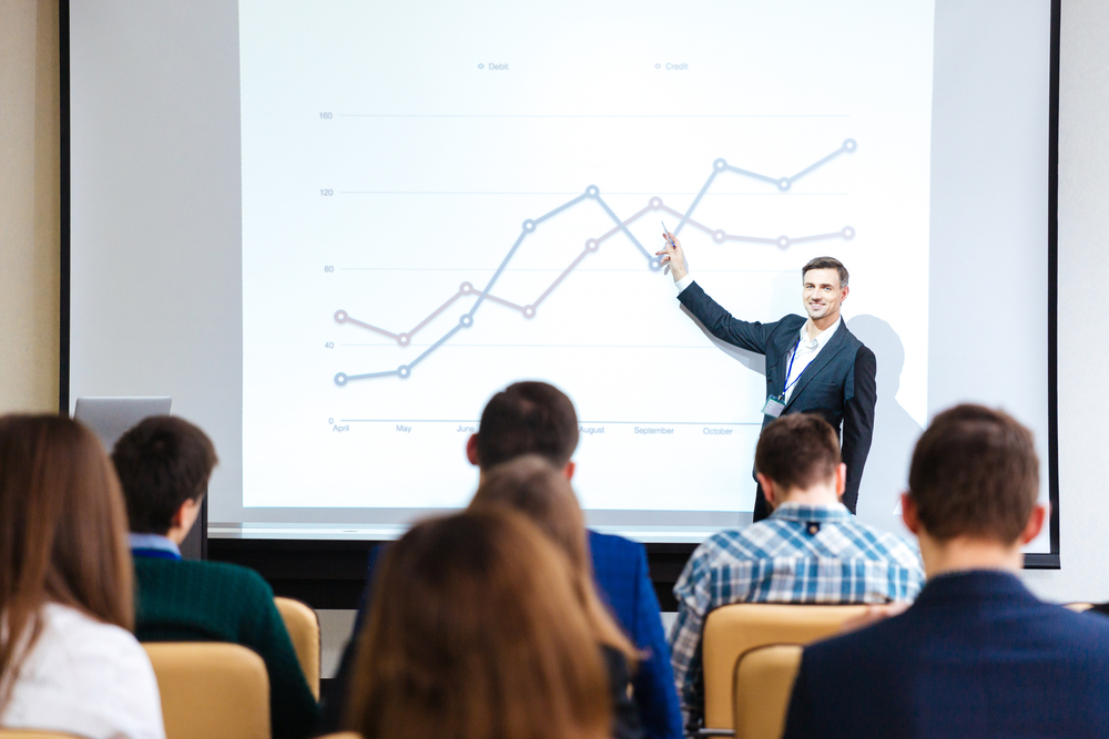 Smiling speaker standing and explaining graphs on business conference in meeting hall